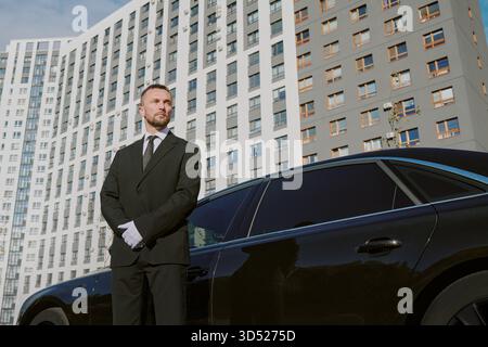 Mann Mittleren Alters, Der Ein Luxusauto Vor Dem Modernen Gebäude Bewacht Stockfoto
