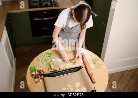Mädchen kneten, rollt, schneidet Kekse aus Teig und legt sie in der Küche auf Backblech. Teenager, der zu Hause Kekse macht, von oben gesehen. Makin Stockfoto