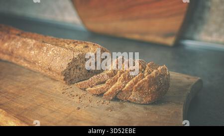 Frisch gebackenes Baguette aus dunklem Brot auf einem Schneidebrett aus Holz, fotografiert auf einer sauberen Tischoberfläche bei weichem Umgebungslicht Stockfoto