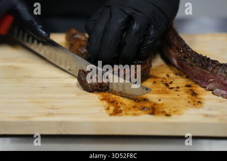 Nahaufnahme eines Küchenchefs mit schwarzen Handschuhen, der ein saftiges gegrilltes Steak auf einem hölzernen Schneidebrett schneidet. Stockfoto