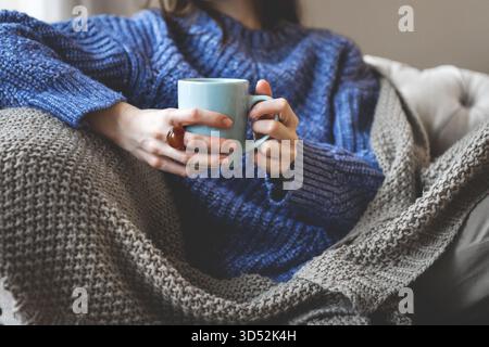 Schließen Sie weibliche Hände, die eine blaue Tasse Tee oder Kaffee halten. Frau in Strickpullover und gemütlichem Karomuster, die sich zu Hause auf dem Sofa entspannt, warm und friedlich drinnen Stockfoto