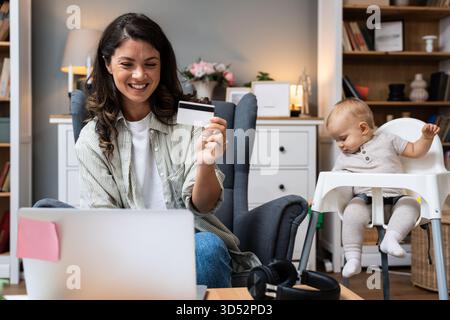Die mobil arbeitende Mutter bestätigt die Online-Zahlung per Video-Chat lächelnd, während sie die Kreditkarte an den Laptop hält, während ihr Baby in einem Hochstuhl sitzt und sich ausbalanciert Stockfoto