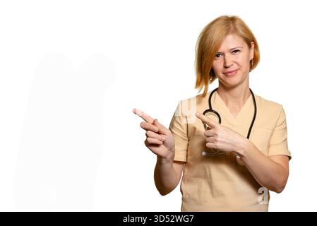 Eine Ärztin, eine Ärztin, zeigt ihre Zeigefinger zur Seite. Sie trägt eine beige Uniform und sieht ernsthaft in die Kamera. Weißes bac Stockfoto