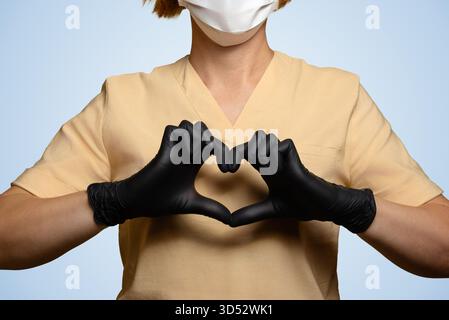 Eine Ärztin, eine Angestellte im Gesundheitswesen. Nahaufnahme der Finger beider Hände, die ein Herz in Brusthöhe bilden. Ihre Hände sind in schwarzen Handschuhen. Sie ist getragen Stockfoto