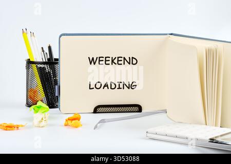 Ein geöffnetes Notizbuch zeigt den Satz WEEKEND LOADING an. In der Nähe befinden sich Schreibgeräte, ein Laptop und weggeworfene, zerknitterte Papiere, alle gegen ein Uni gestellt Stockfoto