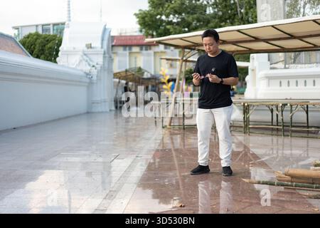 Ein Mann steht in einem Tempelhof in Bangkok und nimmt seine Brille aus dem Etui, während er sich darauf vorbereitet, sie in der ruhigen, traditionellen Umgebung anzuziehen. Stockfoto