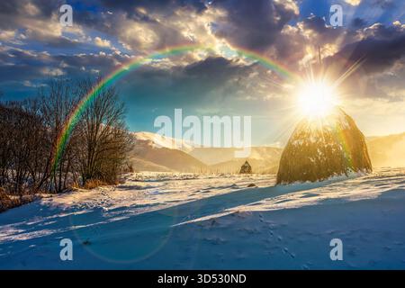 Ländliche Landschaft mit Heuhaufen im Winter bei Sonnenuntergang. Landschaft mit schneebedeckten Hügeln in Bergen im Abendlicht. Niedriger Blickwinkel mit Schnee Stockfoto