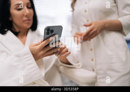 Nahaufnahme einer Frau mittleren Alters und einer Kosmetikerin, die sich an einer Konsultation beteiligt, und der Kunde hält ein Smartphone, um visuelle Informationen über Ästhetik zu erhalten Stockfoto