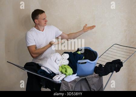 Junger Mann, der die Handfläche neben Wäscheständer und Korb hebt (55 Zeichen). Ein junger Mann hebt seine Hand mit der Handfläche neben einem Wäscheständer mit Kleidung Stockfoto
