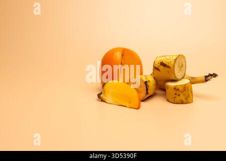 In Scheiben geschnittene Persimmon- und geschnittene Bananenstücke sind auf einer warmen beigen Oberfläche angeordnet. Ihre hellen Farben und weichen Texturen schaffen einen natürlichen fres Stockfoto