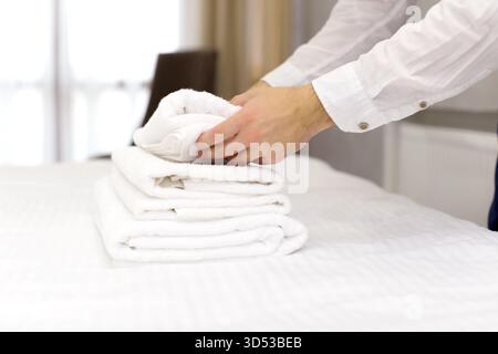 Ordentlich gestapelte weiße Handtücher auf einem Bett mit einer Person, die in einem hellen Zimmer Wäsche faltet Stockfoto