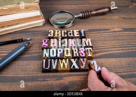 Vollständiges Alphabet aus bunten Vintage-Buchdruckblöcken aus Holz mit Lupe und alten Büchern auf Holzschreibtisch, die Lernpädagogik und Typografie symbolisieren. Stockfoto