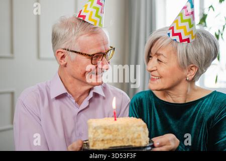 Wünschen Sie sich etwas. Familie Seniorenpaar in Partymütze feiert gemeinsam Geburtstag zu Hause. Ein alter Mann, der Kerzen auf der Geburtstagskuche brennt. Alte Frau wünscht Ehemann alles Gute zum Geburtstag || Model veröffentlicht Stockfoto