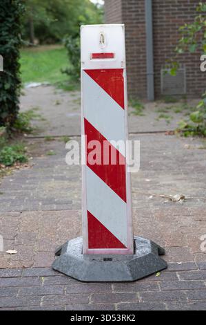 Gestreifter Verkehrspoller auf dem Bürgersteig, verwitterter rot-weißer Pfosten in Betonsockel neben gemauertem Wohnweg, abgenutzte Farbe und Abriebspuren, Stockfoto