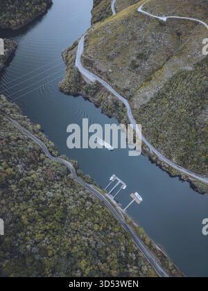 Aus der Vogelperspektive eines Bootes, das durch das dunkle Wasser von Ribeira Sacra in der Nähe der Piers schneidet, mit Straßen, die sich durch die Hügel und grünen Wälder schlängeln, nüchtern, Stockfoto