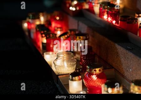 Warme Tribute-Kerzen in roten und weißen Laternen, die entlang der Steinfelsen von Krastmala für den Lāčplēša-Tag angeordnet sind und reflektierende Glasstrukturen hervorheben Stockfoto