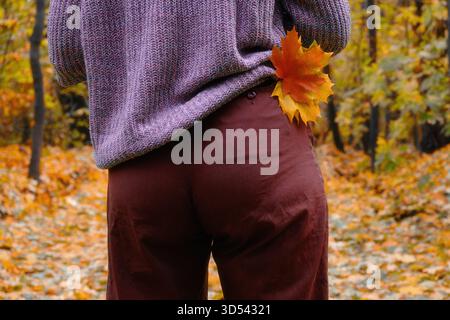 Herbstbild, Rückansicht, ein Mann in einem voluminösen lilafarbenen Pullover und einer braunen Hose hält ein orangenes Ahornblatt in einem Park oder Wald mit gelbem Laub. Stockfoto