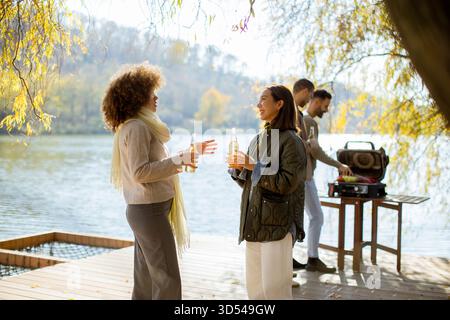 Zwei Freunde genießen erfrischende Getränke, während sie sich auf der Terrasse am See unterhalten. Im Hintergrund wird ein Grill eingerichtet. Stockfoto
