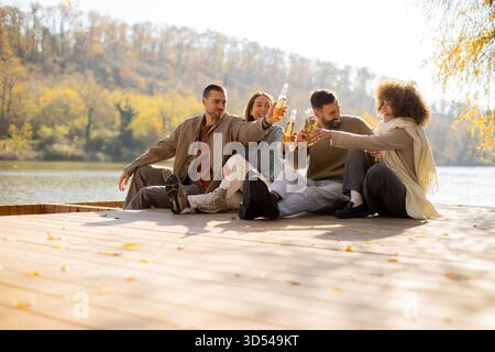 Vier Freunde entspannen sich auf einem hölzernen Dock, während sie an einem friedlichen See, umgeben von lebhaften Herbstlaub, lachen und trinken. Stockfoto