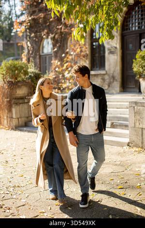 Ein junges Paar geht Arm in Arm auf einem blattbedeckten Weg in einem Park. Das Sonnenlicht filtert durch Bäume, während sie Lächeln und Lachen teilen, umgeben von f Stockfoto