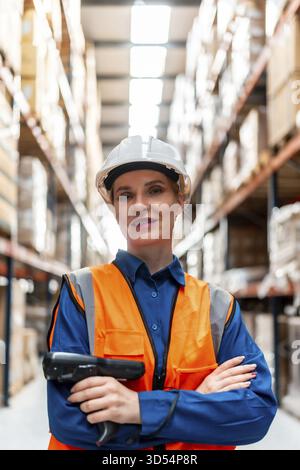 Selbstbewusste Frau, die in einem belebten Logistiklager arbeitet, einen Schutzhelm und eine Sicherheitsweste trägt, einen Barcode-Scanner hält, während sie Lagerbestand und Vorrat verwaltet Stockfoto