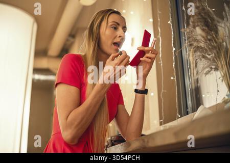 Eine junge Frau in einem roten Kleid trägt Lippenstift mit einem kleinen Spiegel auf. Sie steht an einem Fenster, umgeben von sanften Lichtern und bereitet sich auf ein Abendgeschenk vor Stockfoto