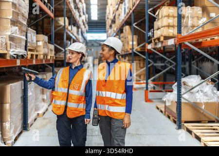 Zwei Frauen, die Schutzhelme und Sicherheitswesten tragen, analysieren den Bestand, indem sie Produktbarcodes in einem modernen Logistiklager scannen Stockfoto