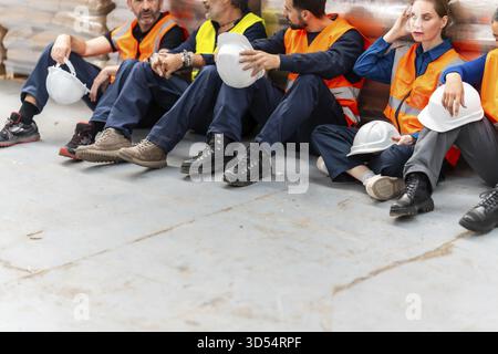 Gruppe von verschiedenen Lagerarbeitern, die auf dem Boden sitzen und eine Pause machen, sich nach einer langen Schicht erschöpft fühlen, Sicherheitskleidung in einem Logistikbereich tragen Stockfoto