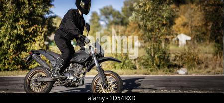 Fahrer auf einem Dirt Bike mit schwarzem Helm und Ausrüstung, mit einer sonnigen Baumkulisse im Freien Stockfoto
