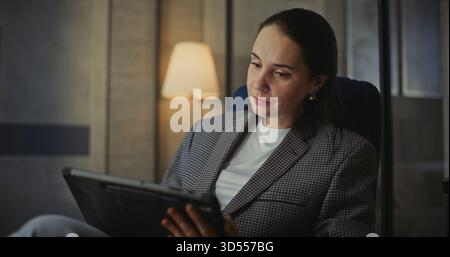 Close-up-Frau mit langen dunklen Haaren sitzt bequem im modernen Hotelsessel und hält Tablet auf Lap. Frau blättert durch Projektdokumente und macht Notizen, wobei sie sich voll und ganz auf die Routine der Remote-Arbeit konzentriert. Stockfoto