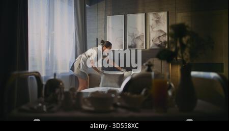 Aufmerksame weibliche Haushälterin in Uniform richtet Bettwäsche und offene Vorhänge in der stilvollen Hotelsuite auf. Konzept Morning Routine des Hotelpersonals, Premium Service, Komfort, Gästeerlebnis. Statische Aufnahme. Stockfoto