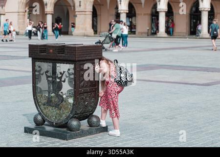 Neugieriges Kind, das in eine dekorative Spendenbox auf dem historischen Hauptmarkt blickt, mit Menschen im Hintergrund. Stockfoto
