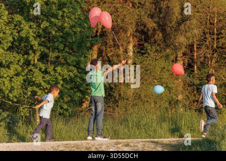 Drei Kinder genießen einen warmen Nachmittag im Park, laufen mit bunten Ballons und haben Spaß in der Sonne. Stockfoto
