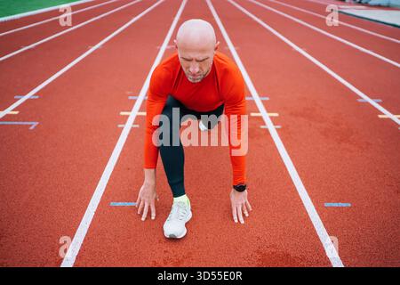 Glatzenläufer mittleren Alters in heller Sportswear dehnt die Beine auf moderner Leichtathletikbahn, zeigt Engagement für Fitness, gesunden Lebensstil und Outdoor-Training Stockfoto