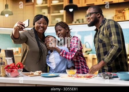 afroamerikanische Mutter macht Fotos mit ihren Kindern und ihrem Mann, die in der Küche kochen, Erinnerungen machen und am Wochenende Spaß haben. Lachen und lustige Zeiten zu Hause. Stockfoto
