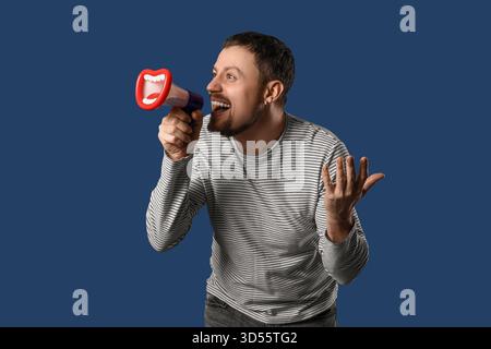 Junger Mann, der in komisches Megaphon auf dunkelblauem Hintergrund schreit Stockfoto