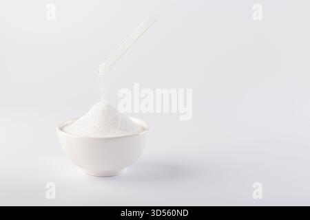 Eine mit weißem Pulver gefüllte Schüssel steht auf einer sauberen Oberfläche. Über dem Pulver befindet sich ein Messlöffel mit feinen Partikeln, die sanft in der Luft abdriften. Stockfoto