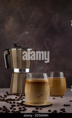 Zwei Gläser Eiskaffee sitzen auf einer glatten Oberfläche, mit einer Kaffeemaschine und verstreuten Kaffeebohnen im Hintergrund, was eine gemütliche Atmosphäre schafft Stockfoto