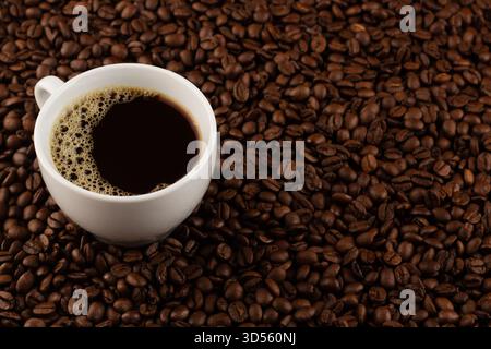 Eine weiße Tasse mit dunklem Kaffee steht auf einer Oberfläche, die mit aromatischen gerösteten Kaffeebohnen bedeckt ist und eine einladende Atmosphäre für Kaffee schafft Stockfoto