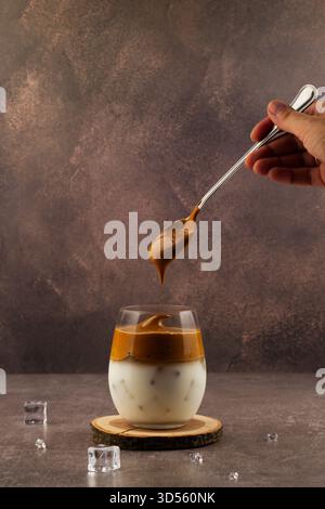 Eine Person hält einen Löffel geschlagenen Kaffee über einem mit Milch und Eis gefüllten Glas. Der cremige Schaum auf der Oberseite sieht einladend aus, perfekt für einen erfrischenden dr. Stockfoto