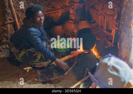 Gasura Ruanda - 8. September 2025; authentisches afrikanisches Dorfleben Frau kocht über offenem Feuer hockend auf dem Boden einer kleinen dunklen Hütte kocht auf offener Tanne Stockfoto