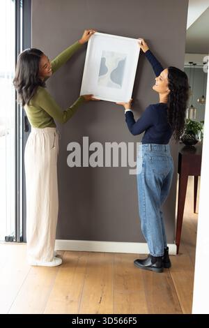 Verschiedene weibliche Freunde hängen abstrakte Kunstdrucke an dunkelgrauer Akzentwand mit Konsolentisch Stockfoto