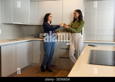 Lächelnde, vielfältige Freundinnen, die an einem kleinen Glasgefäß an der modernen Küchentheke vorbeikommen, mit Smartphone Stockfoto