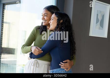 Verschiedene weibliche Freunde, die sich in Atelier in der Nähe eines großen Fensters umarmen, einschließlich abstrakter Malerei Stockfoto