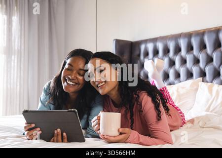 Verschiedene Freundinnen liegen auf dem Bett im Schlafzimmer, verwenden ein Tablet und schlürfen aus weißem Keramikbecher Stockfoto