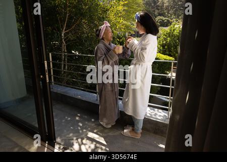 Asiatische Erwachsene Mutter und Tochter unterhalten sich auf dem Balkon im üppigen Garten, halten Tassen und lächeln warm Stockfoto