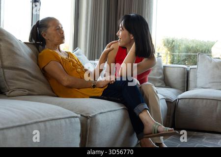 Asiatische Erwachsene Mutter und Tochter plaudern ruhig in einem gemütlichen Wohnzimmer auf der Couch in der Nähe von sonnendurchfluteten Fenstern Stockfoto