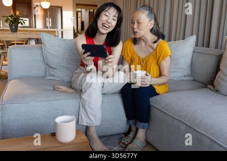 Asiatische ältere Mutter und Tochter sitzen auf dem Sofa, genießen Smartphone und Kaffee zusammen Stockfoto