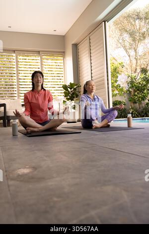 Zwei asiatische Frauen meditieren auf Yogamatten am Pool und genießen friedliche Entspannung. Meditation, Wellness, Ruhe, Achtsamkeit, Zen, Spiritualität Stockfoto