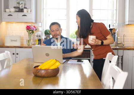 Reifes Ehepaar in der Küche, das über die Arbeit spricht, während er einen Laptop benutzt und Kaffee hält Stockfoto